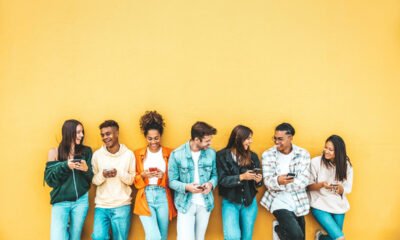 A group of people leaning against a yellow wall, holding and looking at their cell phones.